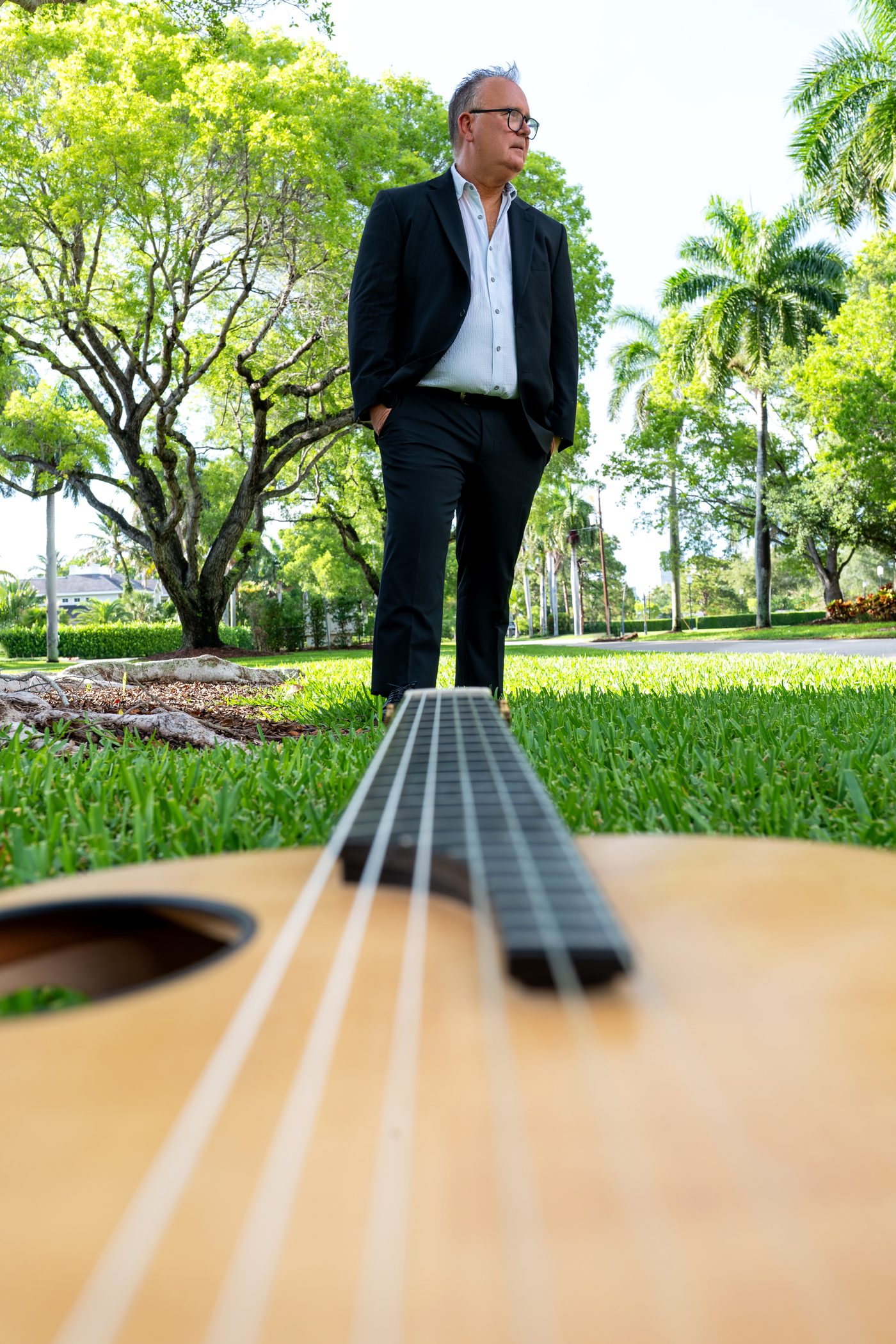 Mark Welch in a dark suit standing on a sunlit lawn, a guitar resting in the foreground